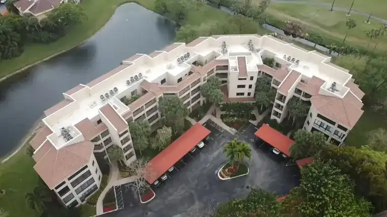 White flat roofs and pink tile edges on a curved condominium building above a palm-lined entrance in La Paz, Boca Raton