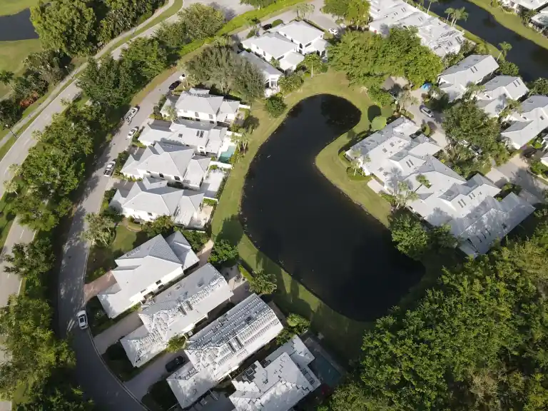 High aerial view of pale tile roofs around a dark curving lake bordered by dense trees in Waters Edge, Boca Raton