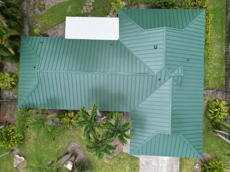 Drone view of a green standing seam metal roof with a white flat-roof section on a house beside a lawn and palms in Boynton Beach
