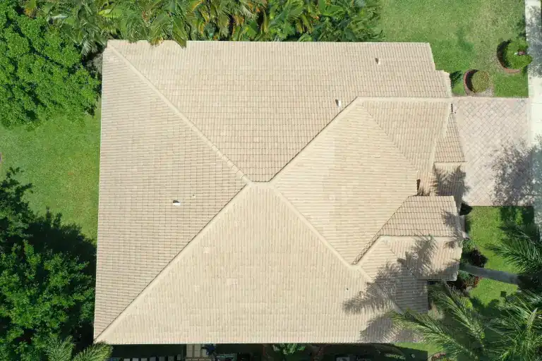 Top-down drone view of a pale sand-colored flat tile hip roof on a single-story house framed by lawn and palms in Boynton Beach