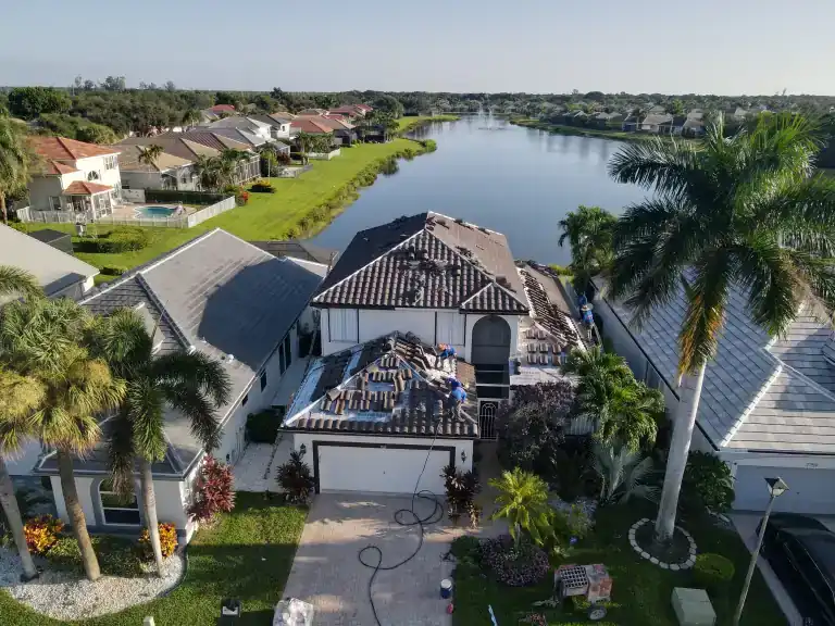 Roof tile installation on a two-story white house with a wide lake and neighboring homes behind in Delray Beach, photographed August 2026