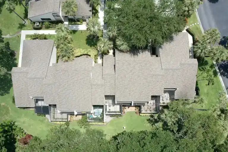 Overhead view of pale shingle roofs with staggered rear patios above a lawn bordered by dense trees in Juno Beach