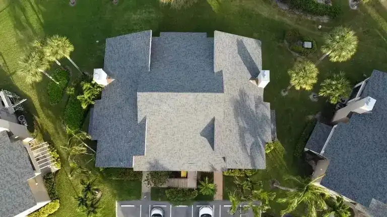 Straight-down view of a gray shingle roof with raised gables and two white chimneys between lawns and a parking row in Jupiter