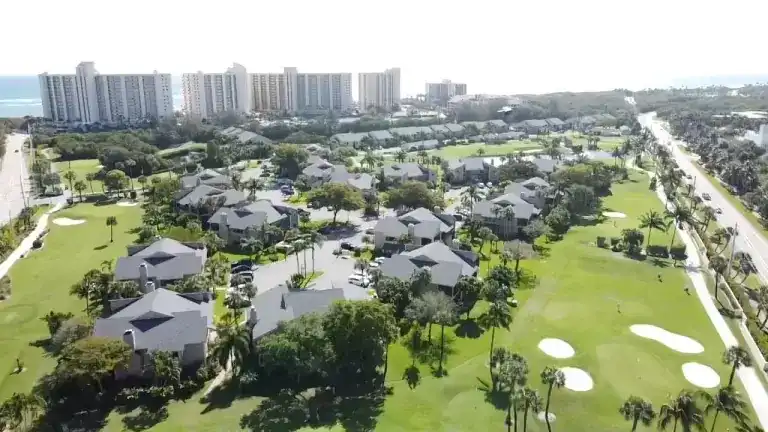 Wide aerial view of gray shingle roofs beside a golf course with sand bunkers and tall coastal buildings beyond in Jupiter