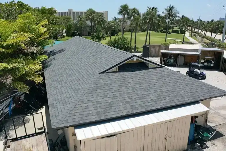 Rear view of a gray shingle roof above a wood-sided building, with golf carts and palms beside a paved yard in Jupiter