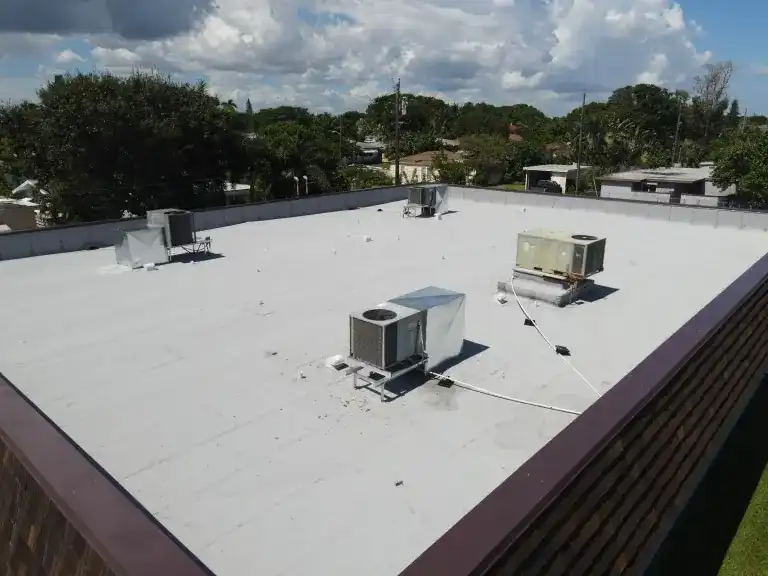 Oblique view across a white flat roof with several air conditioners, a dark parapet and trees beyond in Lake Worth Beach, photographed October 2024