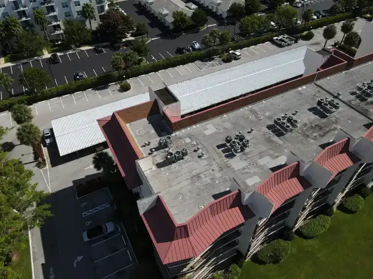 Drone view of a commercial building with a red standing seam metal mansard roof and a white flat roof with rooftop units beside a parking lot in Lantana