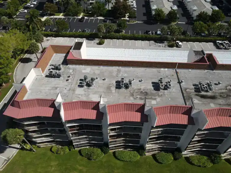Wide drone view of a two-story commercial building with red metal mansard roofing and a white flat roof over a parking lot in Lantana