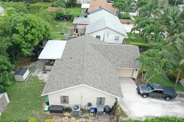 Angled drone view of a gray shingle roof on a single-story house with a carport, driveway and palms in Lantana