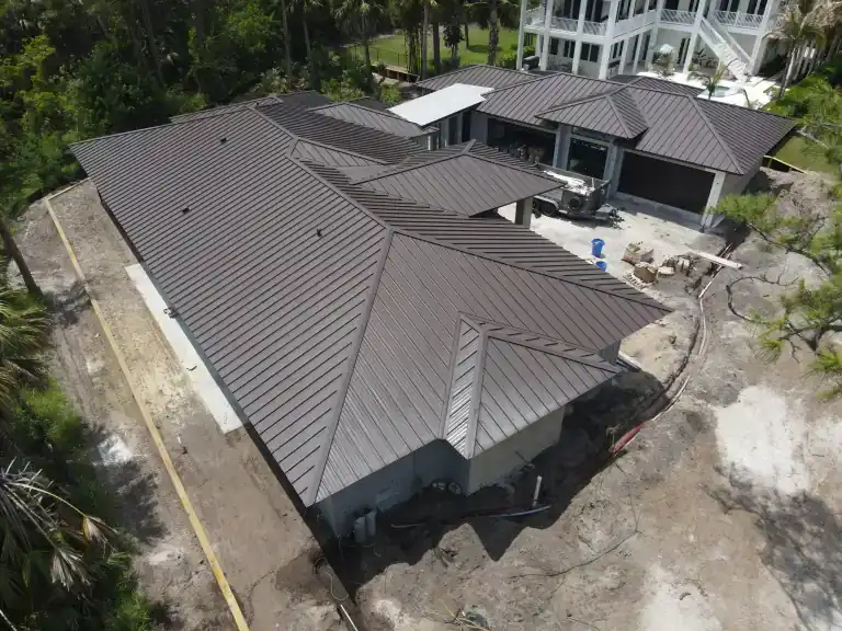 Techo oscuro de metal con juntas alzadas sobre una casa nueva con patio abierto, materiales de construcción y árboles detrás, fotografiado en mayo de 2026