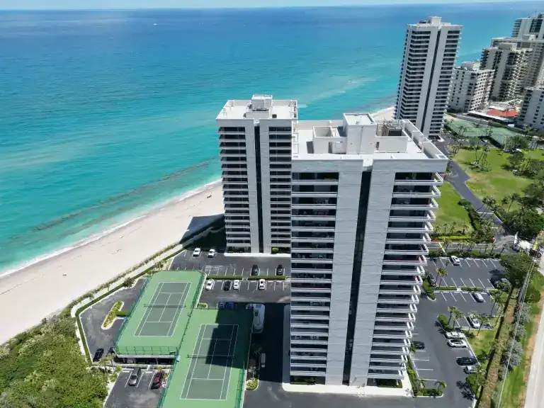 Ocean-facing view of white flat-roofed condominium towers above green tennis courts and a parking lot in Riviera Beach, photographed April 2024