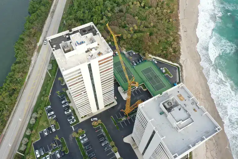 Steep aerial view of two tower roofs and a crane between a coastal road and the beach in Riviera Beach