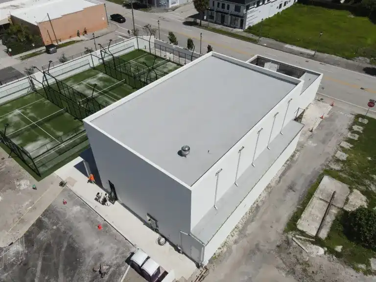 Gray sports building with a white flat roof, two green courts behind and an unpaved area beside it in West Palm Beach, photographed October 2024