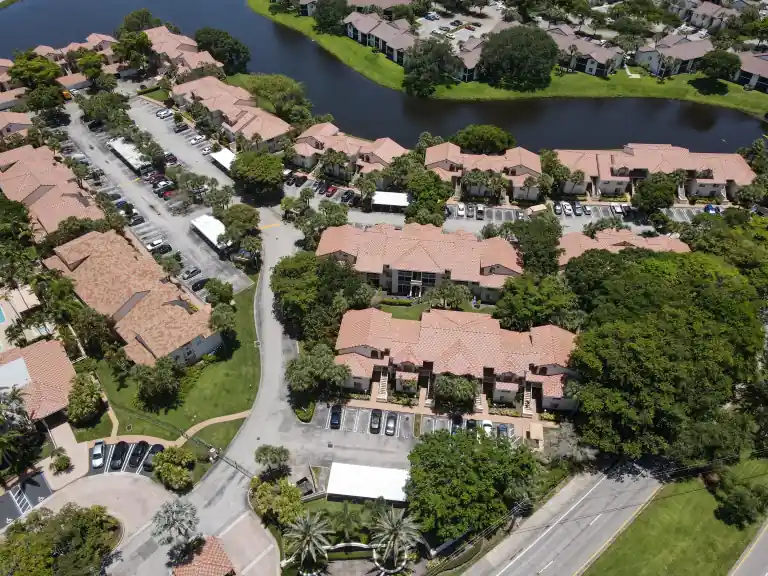 Aerial view of pink tile apartment roofs and winding parking lanes beside a tree-lined lake in Sterling Villages, West Palm Beach, completed June 2026