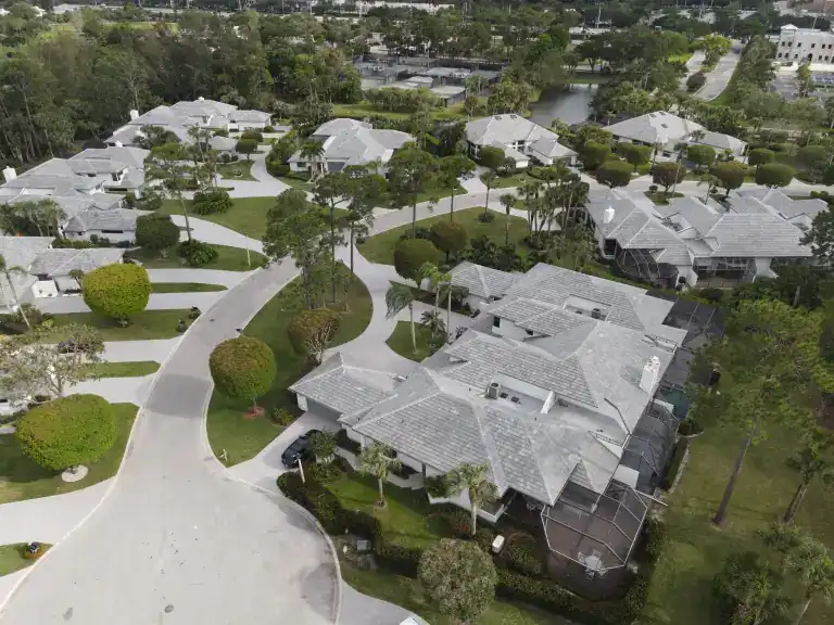Pale tile roofs along a curving residential street with shaped hedges and palms in West Palm Beach, photographed April 2024