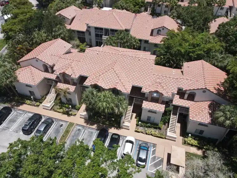 Pink tile roofs on two low apartment buildings behind parked cars and mature shade trees in Sterling Villages, West Palm Beach, completed June 2026